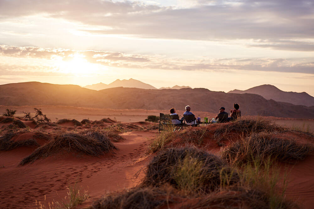 Dieses Bild zeigt unsere Gruppe beim Sundowner in den Hügeln des NamibRand im weichen Abendlicht mit untergehender Sonne.