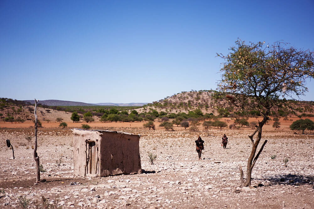 The picture shows an impression of the Himba huts and two Himba walking towards the visitor